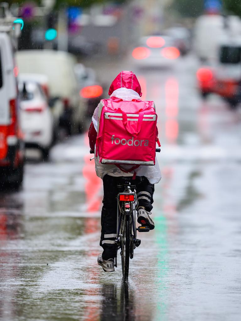 Das Bild zeigt einen Essenslieferanten mit nassem Rucksack, der in der Großstadt zwischen Autos mit einem Fahrrad durch eine regennasse Straße fährt.