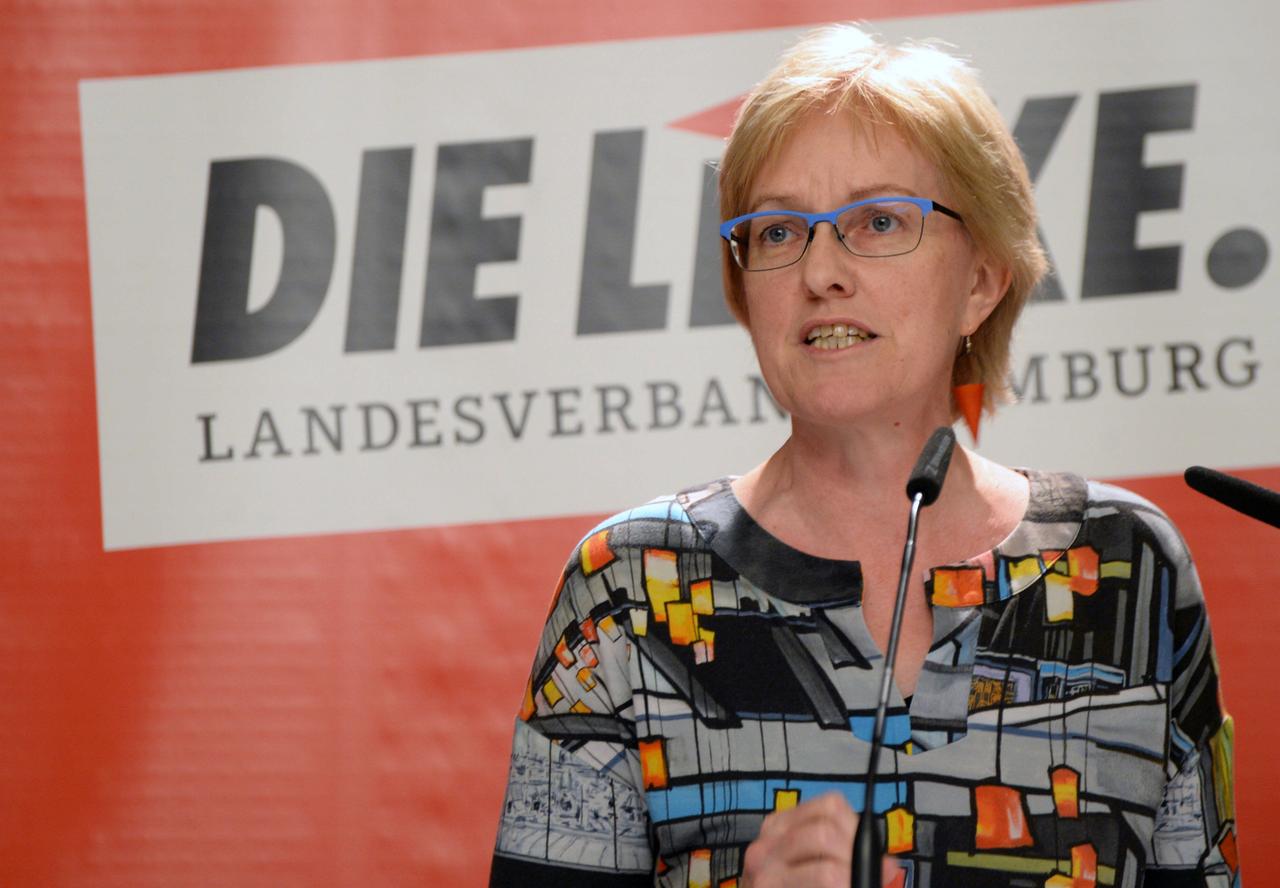 Heike Sudmann steht am Rednerpult, im Hintergrund ist das Logo der Partei "Die Linke" zu sehen: Sie trägt die Haare kurz, hat eine Brille und eine bunte Bluse.