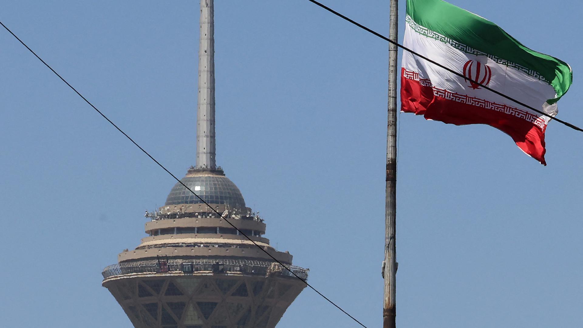 Das Bild zeigt eine wehende iranische Fahne. Im Hintergrund steht ein Fernsehturm der Hauptstadt.