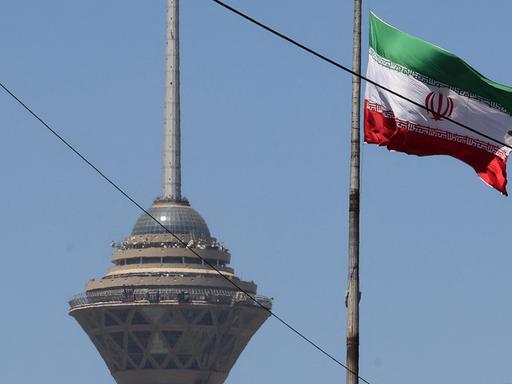 Das Bild zeigt eine wehende iranische Fahne. Im Hintergrund steht ein Fernsehturm der Hauptstadt.
