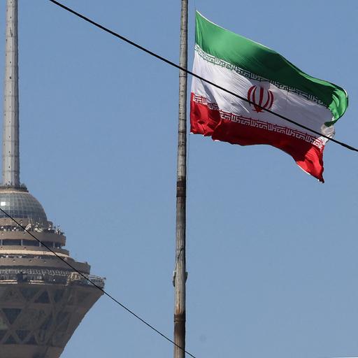 Das Bild zeigt eine wehende iranische Fahne. Im Hintergrund steht ein Fernsehturm der Hauptstadt.