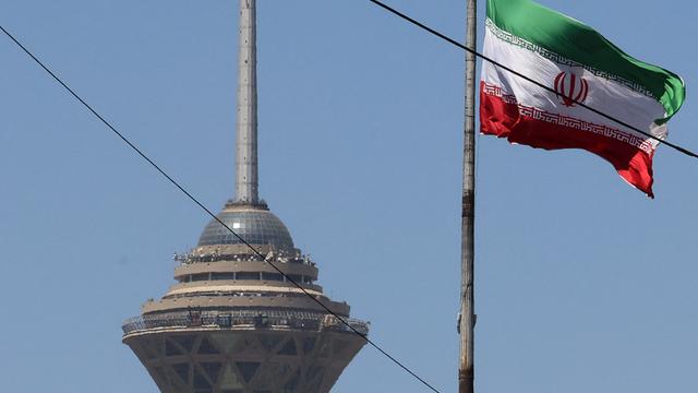 Das Bild zeigt eine wehende iranische Fahne. Im Hintergrund steht ein Fernsehturm der Hauptstadt.