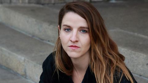 Helene Hegemann sind auf den Stufen vor einem Gebäude und schaut in die Kamera. Sie trägt ein schwarzes Oberteil, ihre langen Haare sind braun mit helleren Strähnen.