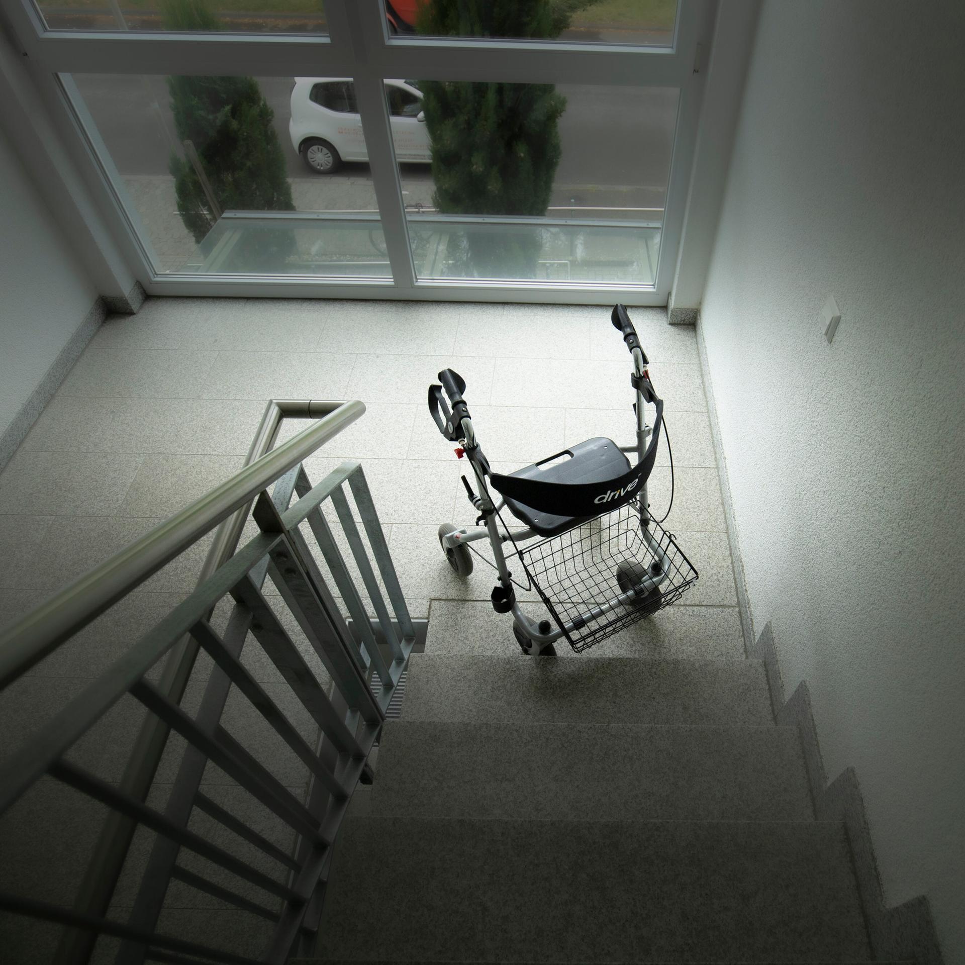Ein Rollator steht am unteren Ende einer Treppe. Das Bild ist dunkel gehalten.