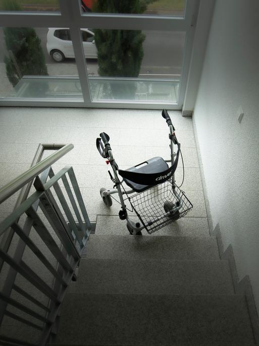 Ein Rollator steht am unteren Ende einer Treppe. Das Bild ist dunkel gehalten.