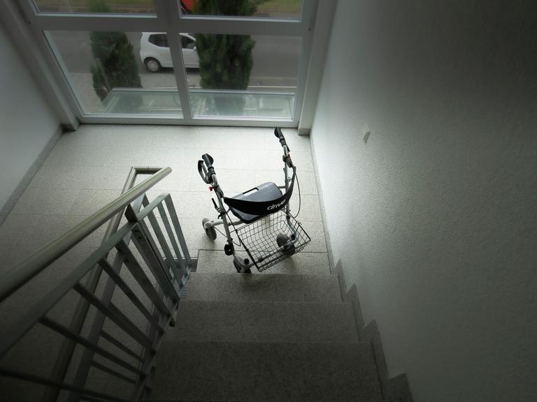 Ein Rollator steht am unteren Ende einer Treppe. Das Bild ist dunkel gehalten.