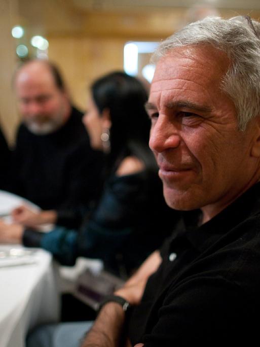 Der verstorbene Investmentbanker und verurteilte Sexualstraftäter Jeffrey Epstein im Porträt an einem gedeckten Tisch mit Personen im Hintergrund. 