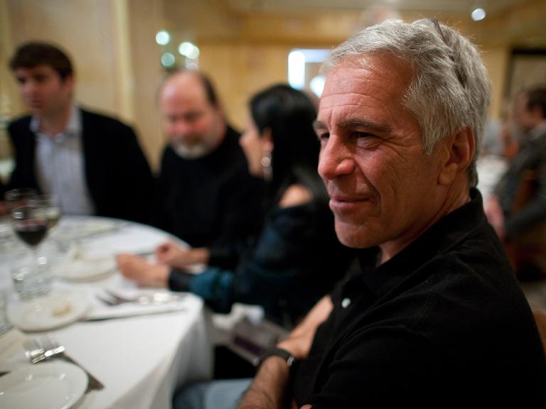 Der verstorbene Investmentbanker und verurteilte Sexualstraftäter Jeffrey Epstein im Porträt an einem gedeckten Tisch mit Personen im Hintergrund. 