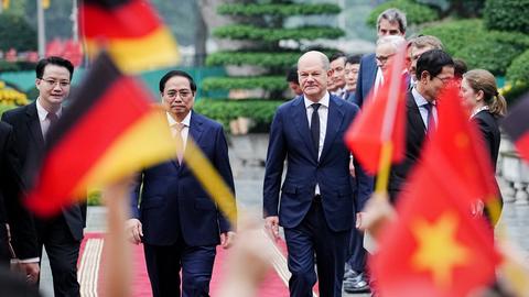 Bundeskanzler Olaf Scholz auf Staatsbesuch in Hanoi, neben ihm der vietnamesische Premier Phan Minh Chinh. Zu beiden Seiten des roten Teppichs schwenken Menschen deutsche und vietnamesische Fahnen.