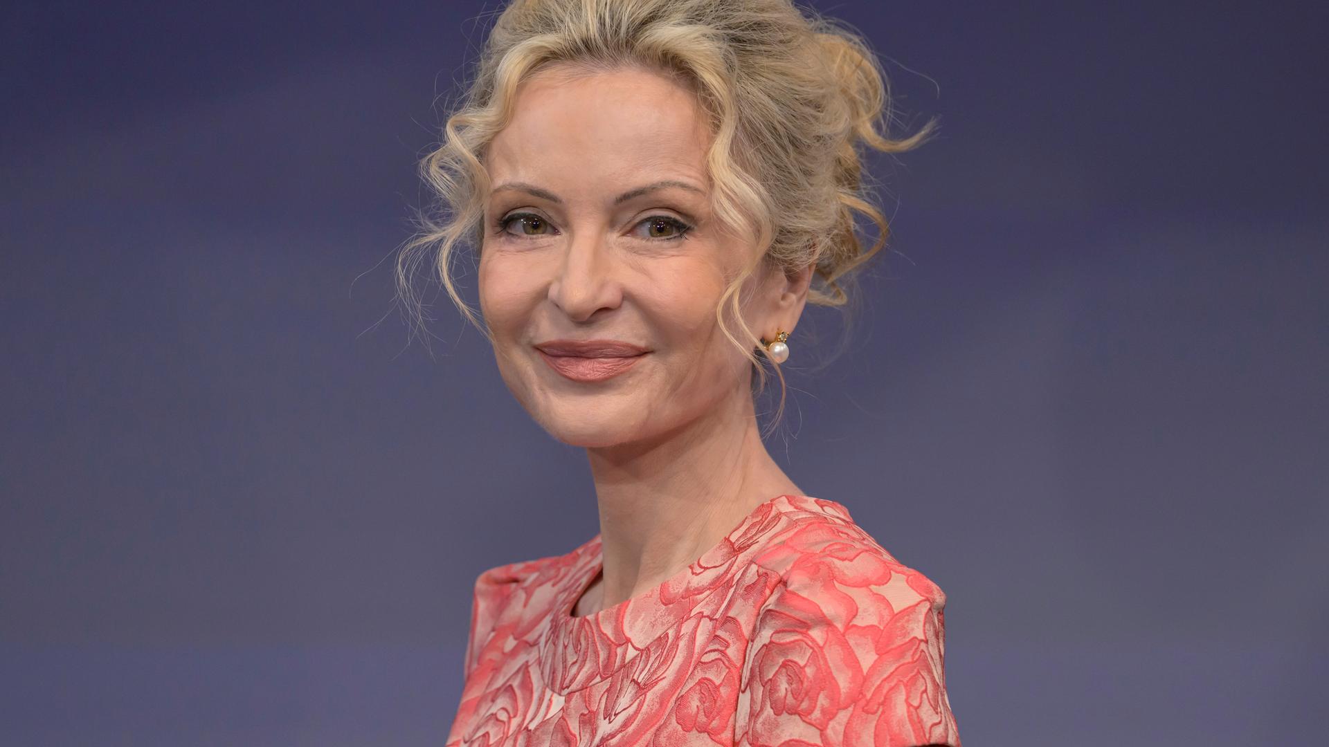 Sandra Navidi sitzt vor einem blauen Hintergrund in einem Fernsehstudio, trägt eine lachsfarbene Bluse und lächelt in die Kamera