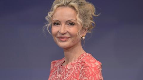 Sandra Navidi sitzt vor einem blauen Hintergrund in einem Fernsehstudio, trägt eine lachsfarbene Bluse und lächelt in die Kamera