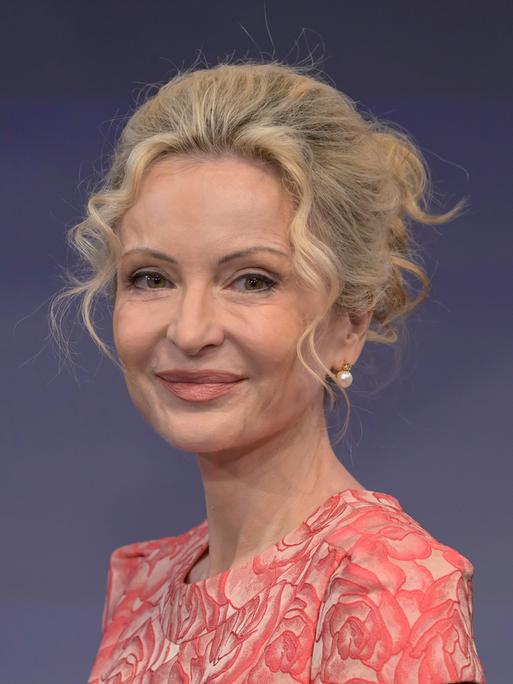 Sandra Navidi sitzt vor einem blauen Hintergrund in einem Fernsehstudio, trägt eine lachsfarbene Bluse und lächelt in die Kamera