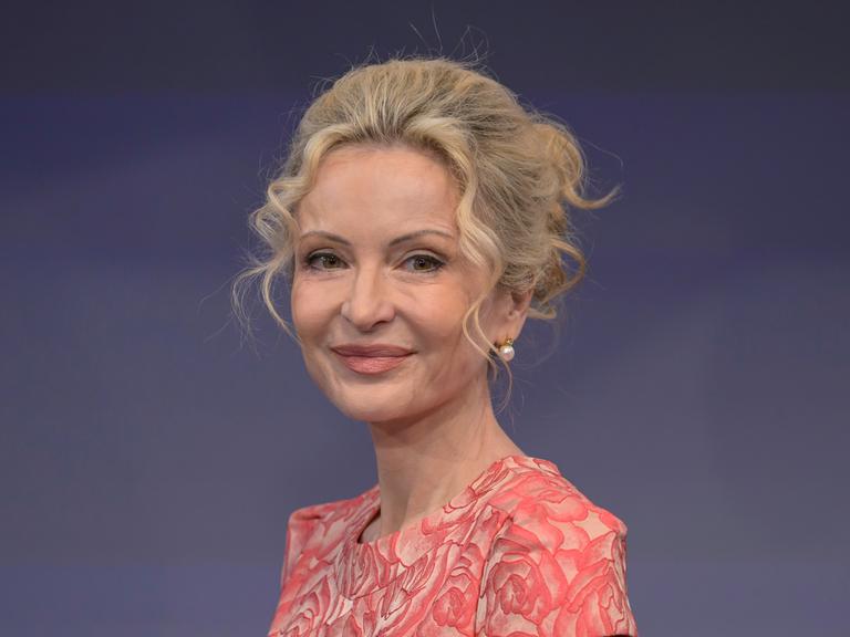 Sandra Navidi sitzt vor einem blauen Hintergrund in einem Fernsehstudio, trägt eine lachsfarbene Bluse und lächelt in die Kamera