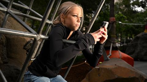 Ein blondes Mädchen sitzt auf einem Klettergerüst und schaut in ihr Smartphone.