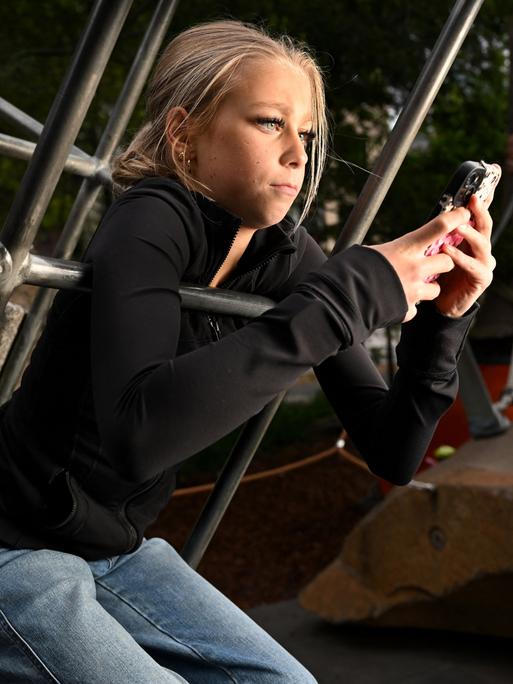 Ein blondes Mädchen sitzt auf einem Klettergerüst und schaut in ihr Smartphone.