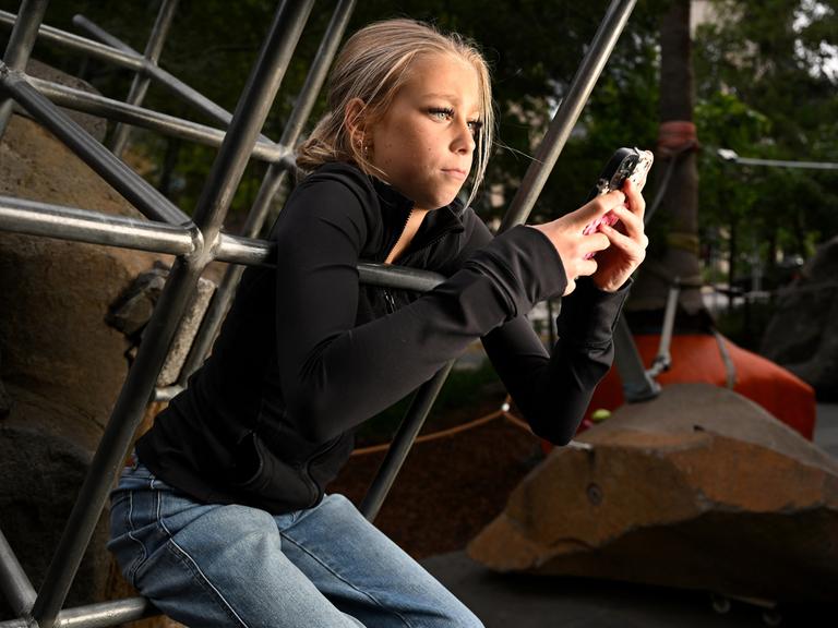 Ein blondes Mädchen sitzt auf einem Klettergerüst und schaut in ihr Smartphone.