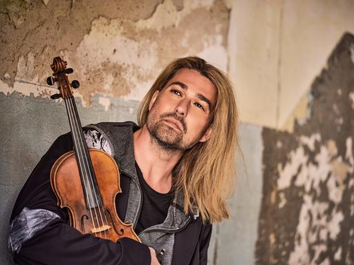 Ein Mann mit blonden, halblangen Haaren, im Arm eine Geige. Er steht in einem alten Gebäude, der Putz blättert ab, er schaut versonnen in die Ferne.