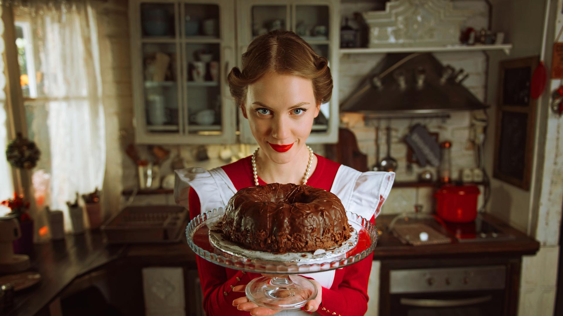 Eine Frau in einem rot-weißen Kleid im Stil der 1950er Jahre und einer eben solchen Frisur, hält einen mit Schokolade glasierten Gugelhupf in die Kamera. Sie trägt eine Perlenkette und schaut leicht von unten in die Kamera. (Symbolbild)