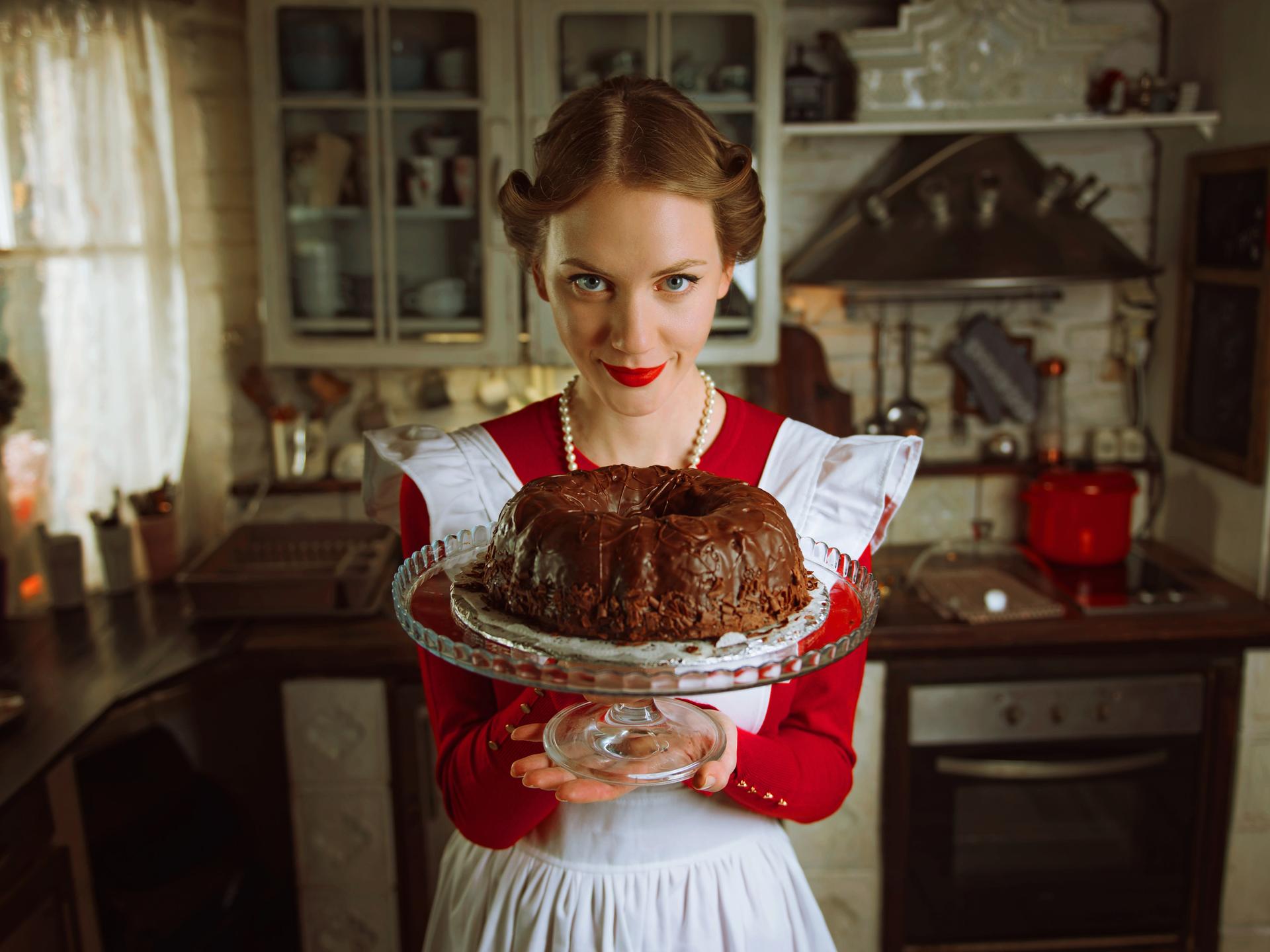 Eine Frau in einem rot-weißen Kleid im Stil der 1950er Jahre und einer eben solchen Frisur, hält einen mit Schokolade glasierten Gugelhupf in die Kamera. Sie trägt eine Perlenkette und schaut leicht von unten in die Kamera. (Symbolbild)