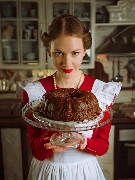 Eine Frau in einem rot-weißen Kleid im Stil der 1950er Jahre und einer eben solchen Frisur, hält einen mit Schokolade glasierten Gugelhupf in die Kamera. Sie trägt eine Perlenkette und schaut leicht von unten in die Kamera. (Symbolbild)
