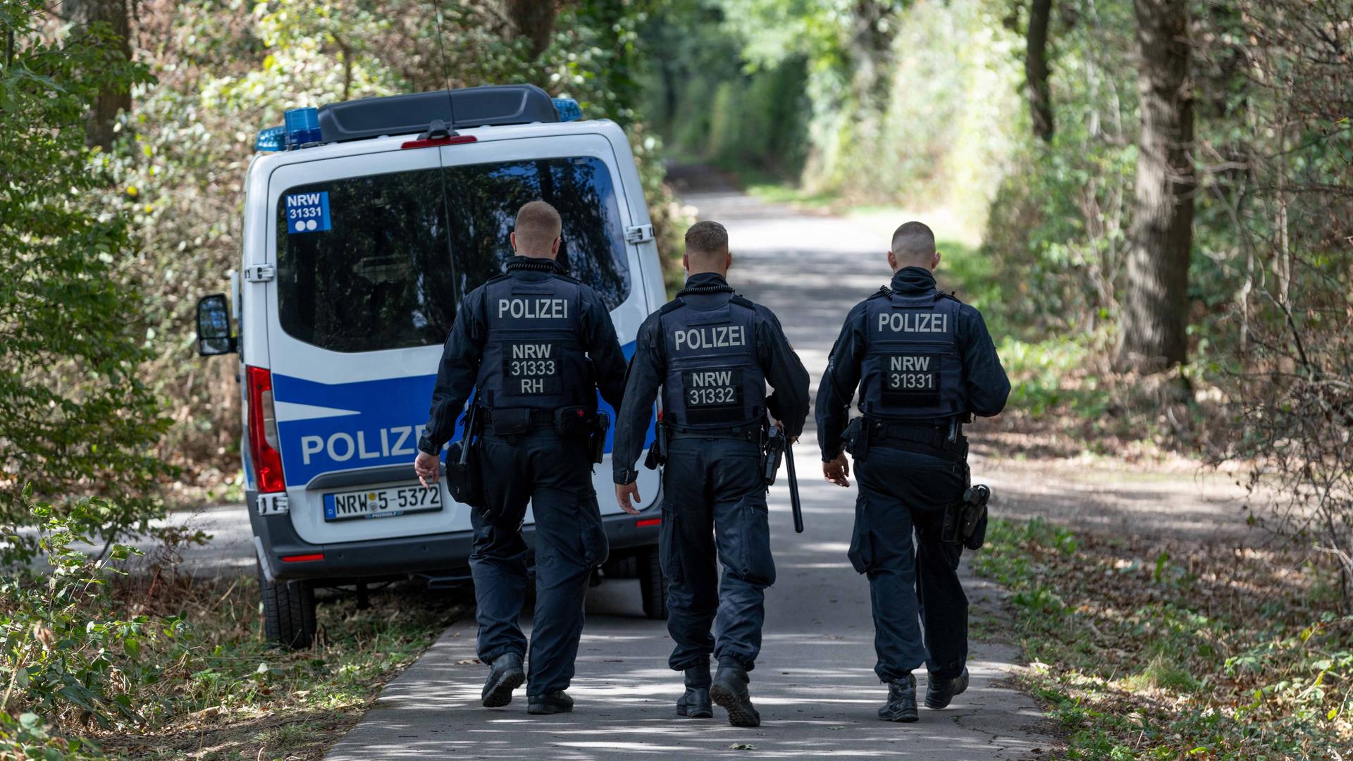 Drei Polizisten laufen auf einer Straße durch einen Wald. Davor steht ein Polizeiwagen. Drei Polizisten laufen auf einer Straße durch einen Wald. Davor steht ein Polizeiwagen.