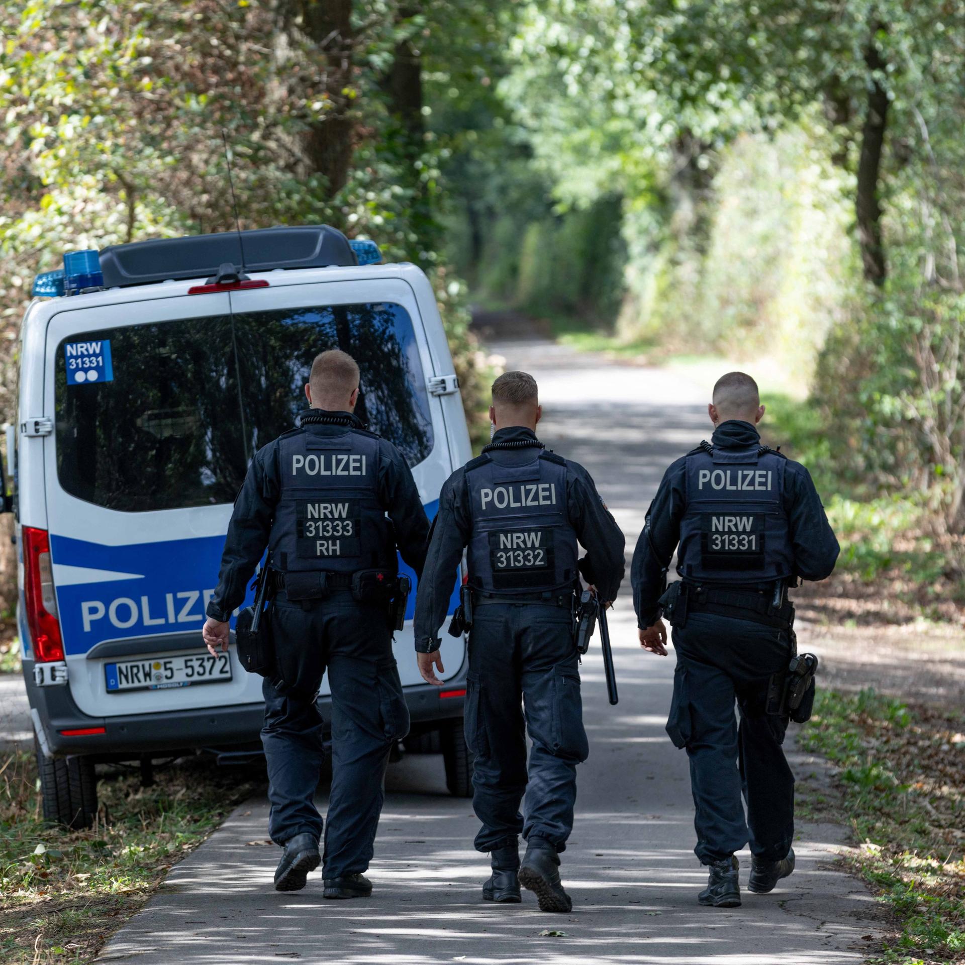 Drei Polizisten laufen auf einer Straße durch einen Wald. Davor steht ein Polizeiwagen.