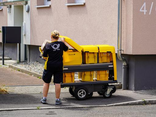 Eine Briefträgerin nimmt Briefe aus ihrem modernen Postwagen der Deutschen Post, um sie in die Briefkästen eines Mehrfamilienhauses einzuwerfen.