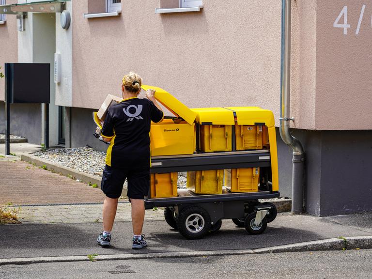 Eine Briefträgerin nimmt Briefe aus ihrem modernen Postwagen der Deutschen Post, um sie in die Briefkästen eines Mehrfamilienhauses einzuwerfen.
