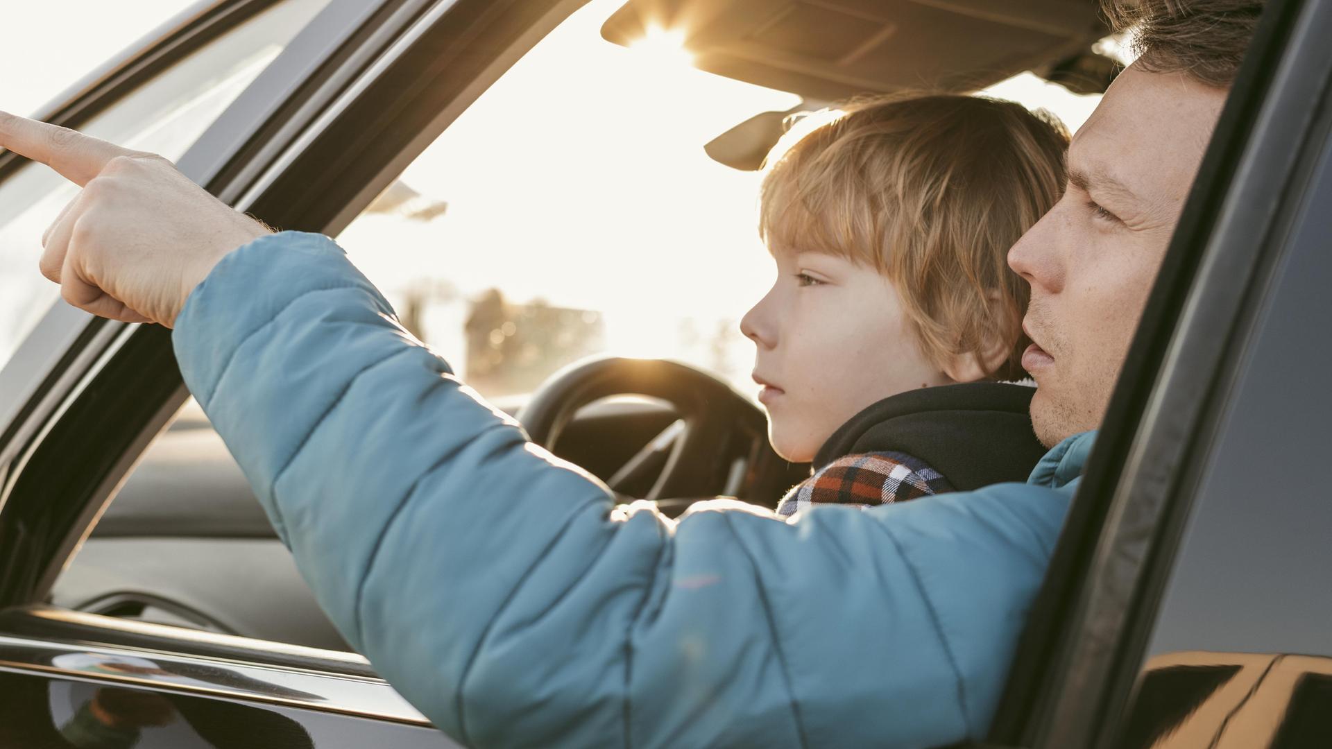 Ein Kind sitzt bei seinem Vater in einem Auto auf dem Schoß. Das Fenster ist geöffnet und der Vater zeigt auf etwas, das Kind blickt hinterher.