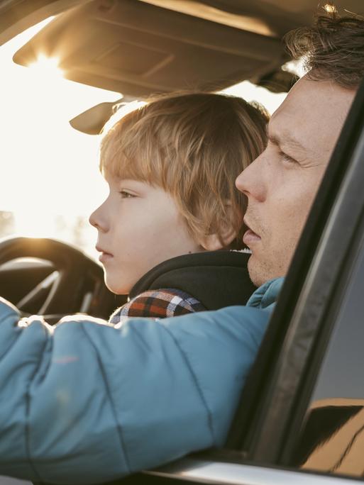 Ein Kind sitzt bei seinem Vater in einem Auto auf dem Schoß. Das Fenster ist geöffnet und der Vater zeigt auf etwas, das Kind blickt hinterher.