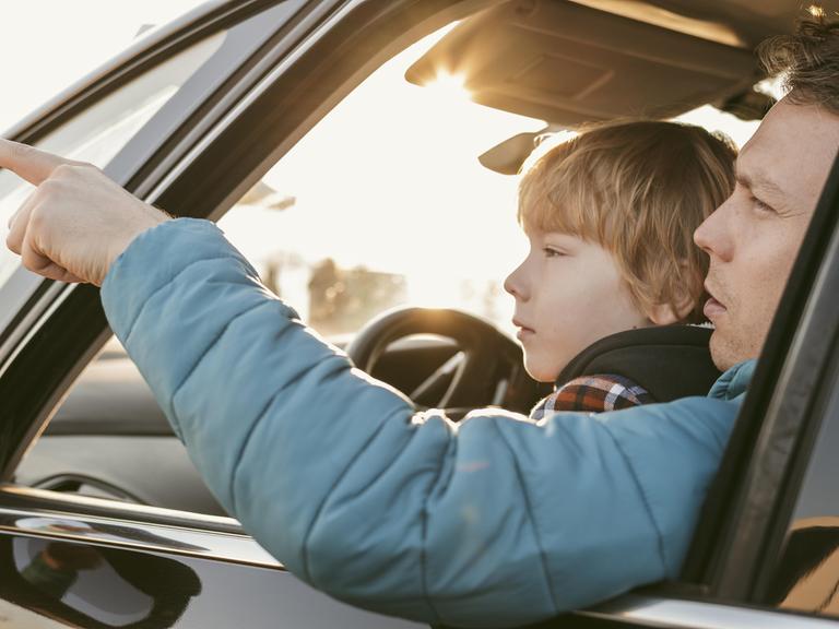 Ein Kind sitzt bei seinem Vater in einem Auto auf dem Schoß. Das Fenster ist geöffnet und der Vater zeigt auf etwas, das Kind blickt hinterher.
