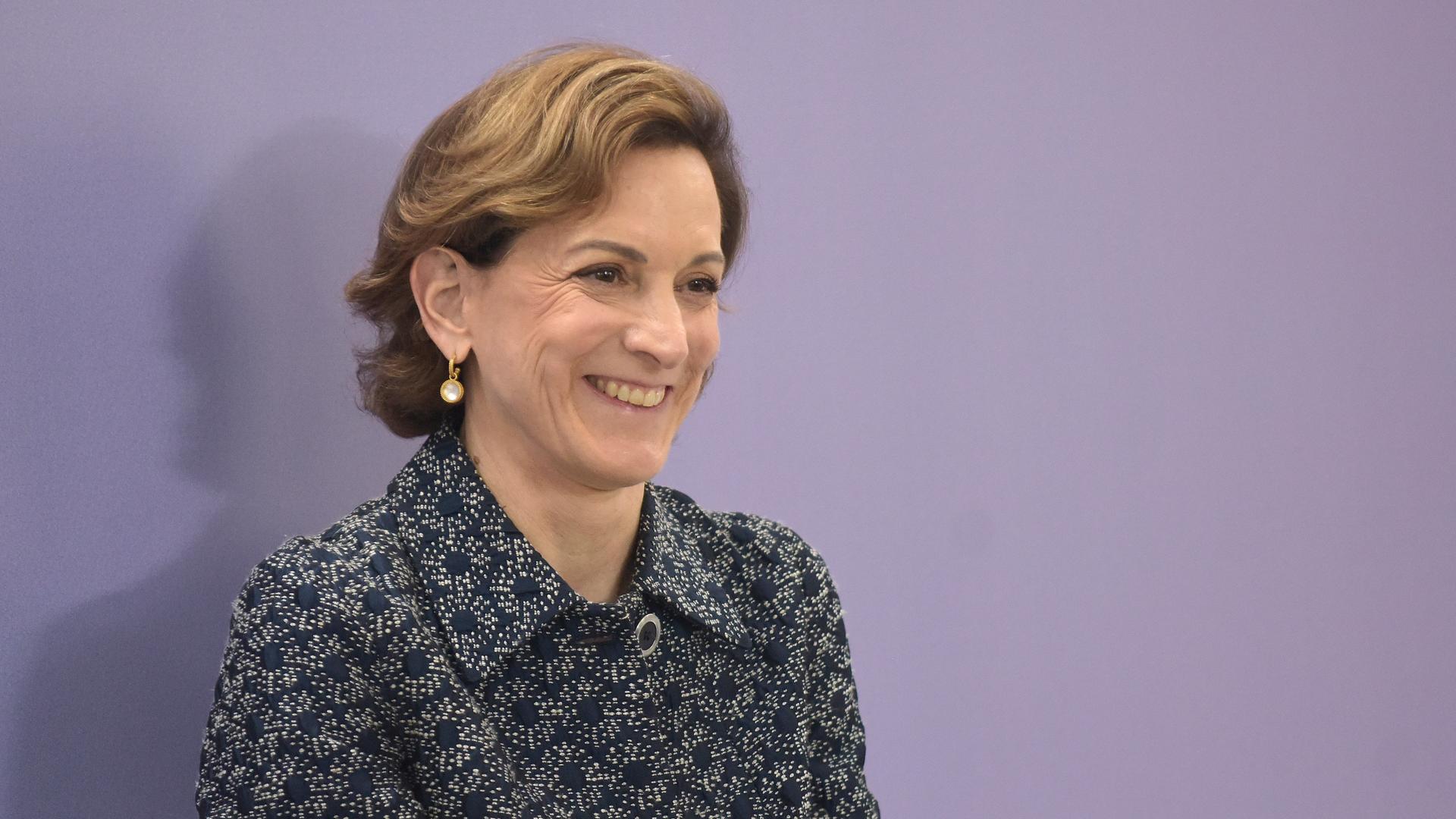 Anne Applebaum trägt eine gemusterte Bluse, steht vor einem violetten Hintergrund und schaut an der Kamera vorbei