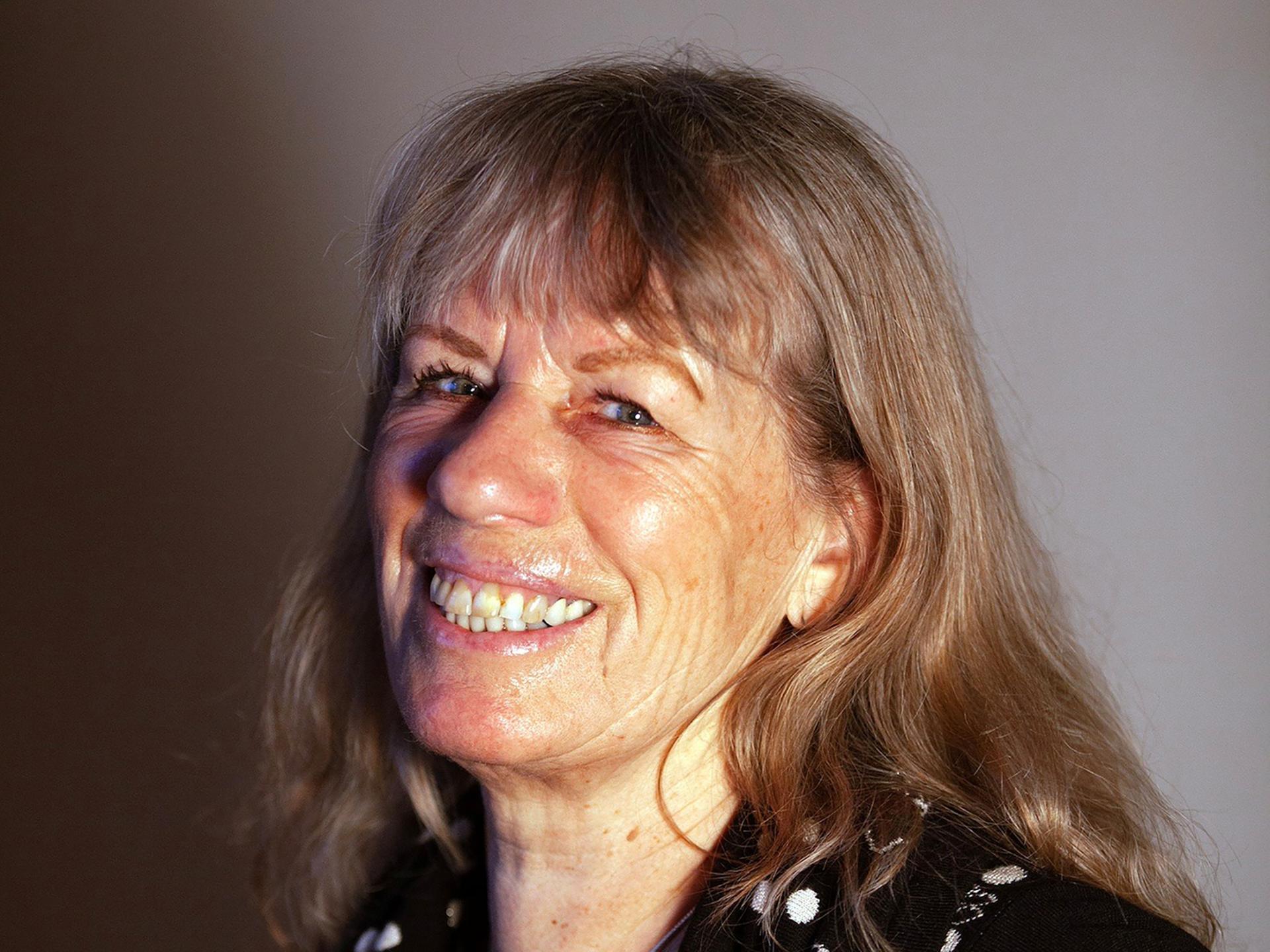 Bettina Kalisch sitzt vor einer grauen Wand und lacht in die Kamera.