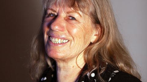 Bettina Kalisch sitzt vor einer grauen Wand und lacht in die Kamera.