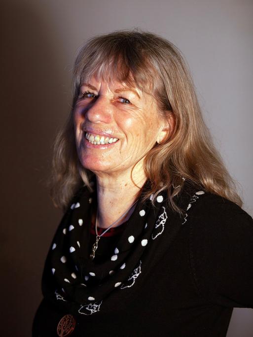 Bettina Kalisch sitzt vor einer grauen Wand und lacht in die Kamera.