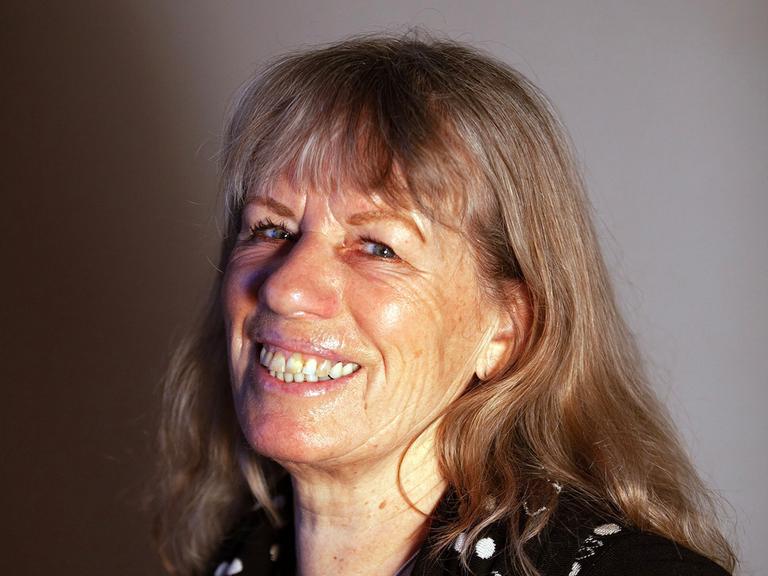 Bettina Kalisch sitzt vor einer grauen Wand und lacht in die Kamera.