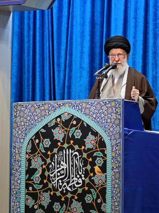 Ayatalloh Ali Khamenei bei Rede vor blauem Samthintergrund neben iranischer Flagge
