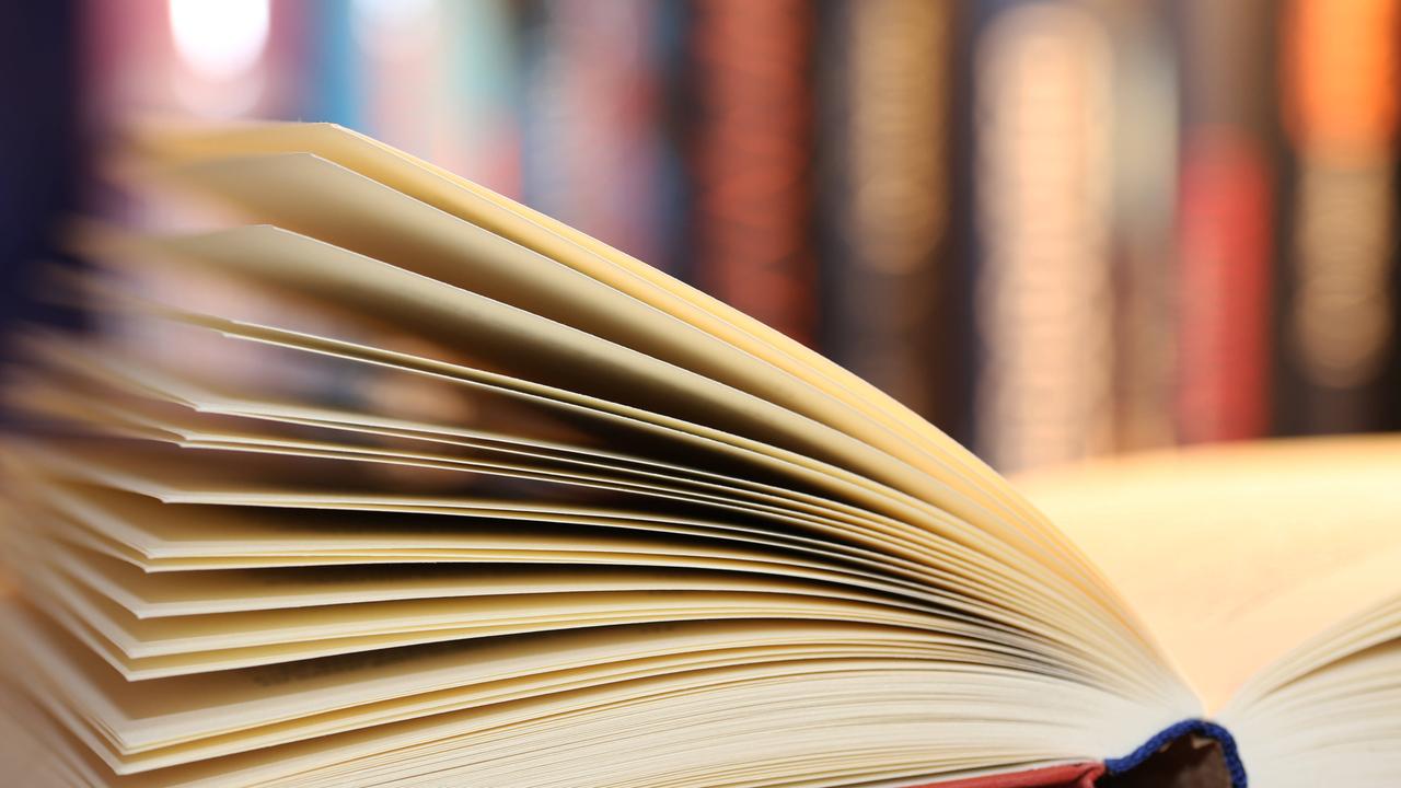 Ein aufgeschlagenes Buch. Sehr unscharf im Hintergrund stehen weitere Bücher in einem Regal 