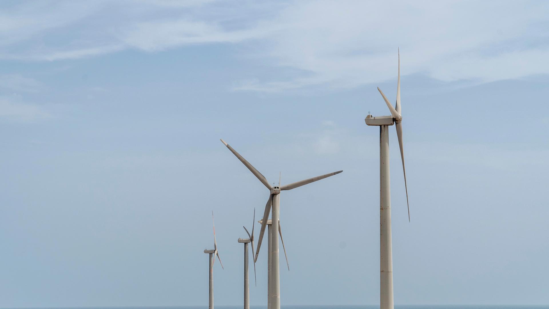 Windräder stehen an der Küste, im Hintergrund ist das Meer zu sehen.