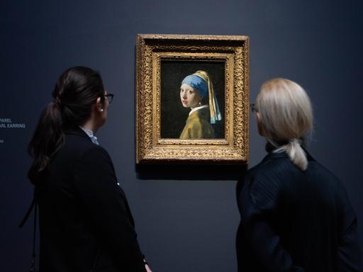 Zwei Frauen stehen vor einem Gemälde und betrachten es. Das Bild hängt an einer dunklen Wand. Es ist das Gemälde "Mädchen mit dem Perlenohrgehänge" des niederländischen Malers Jan Vermeer. Es zeigt ein junge Frau im Halbprofil mit einem hell leuchtenden Perlenohrring.