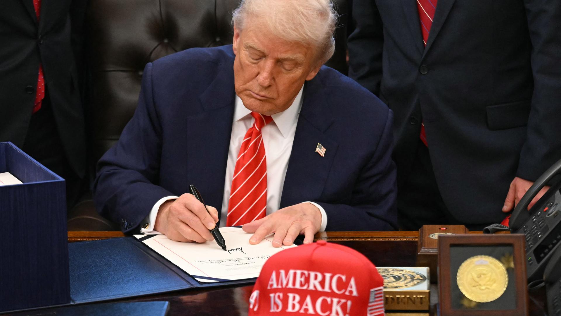 US-Präsident Trump unterschreibt ein Dokument im Weißen Haus. Vor ihm auf dem Schreibtisch eine Baseball-Mütze mit der Aufschrift "America is back" (Amerika ist zurück). Hinter Trump stehen mehrere Personen. US-Präsident Trump unterschreibt ein Dokument im Weißen Haus. Vor ihm auf dem Schreibtisch eine Baseball-Mütze mit der Aufschrift "America is back" (Amerika ist zurück). Hinter Trump stehen mehrere Personen.