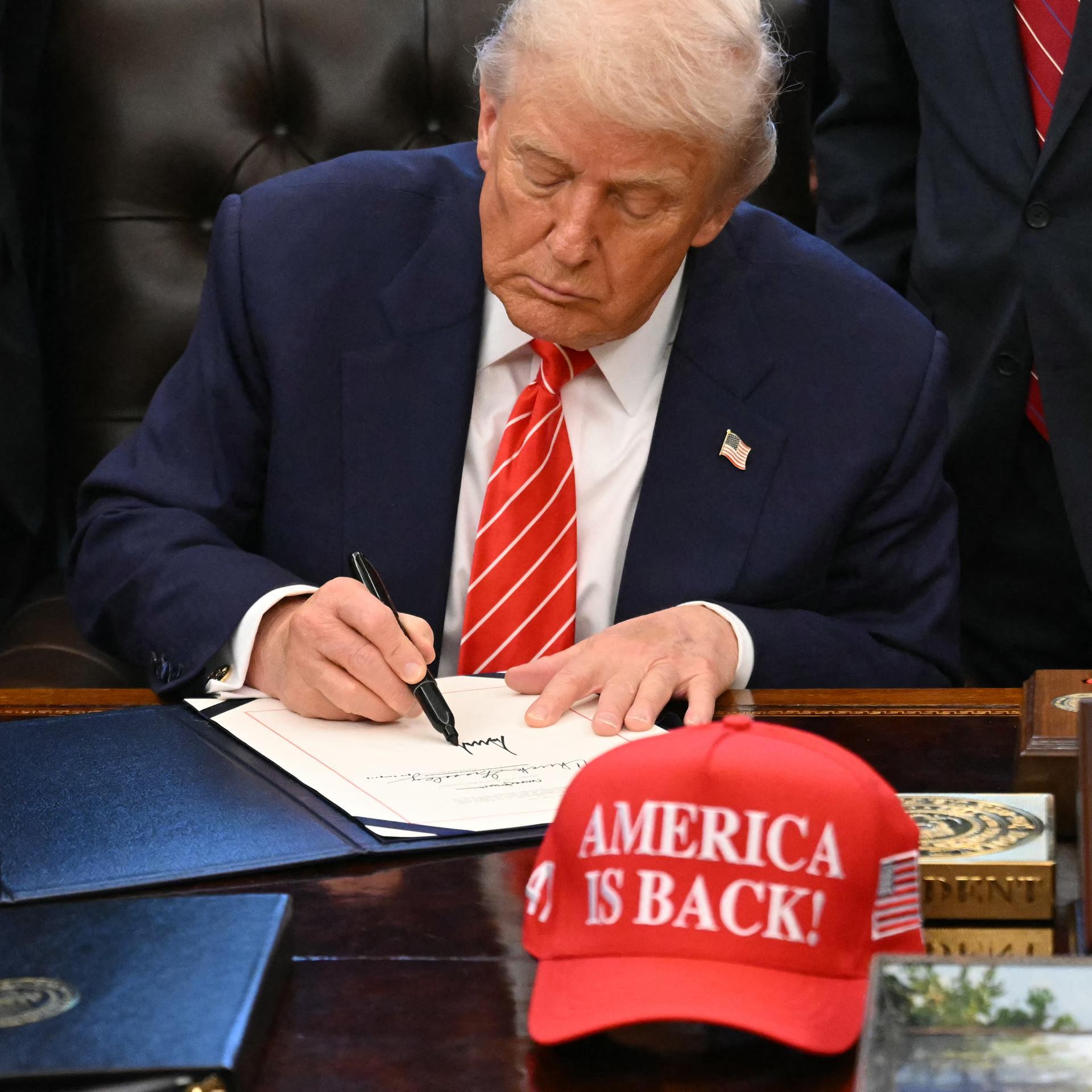 US-Präsident Trump unterschreibt ein Dokument im Weißen Haus. Vor ihm auf dem Schreibtisch eine Baseball-Mütze mit der Aufschrift "America is back" (Amerika ist zurück). Hinter Trump stehen mehrere Personen.
