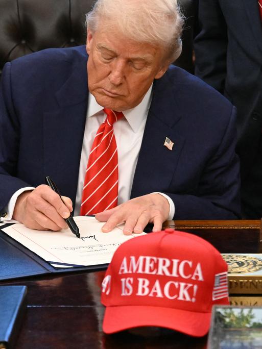 US-Präsident Trump unterschreibt ein Dokument im Weißen Haus. Vor ihm auf dem Schreibtisch eine Baseball-Mütze mit der Aufschrift "America is back" (Amerika ist zurück). Hinter Trump stehen mehrere Personen. US-Präsident Trump unterschreibt ein Dokument im Weißen Haus. Vor ihm auf dem Schreibtisch eine Baseball-Mütze mit der Aufschrift "America is back" (Amerika ist zurück). Hinter Trump stehen mehrere Personen.