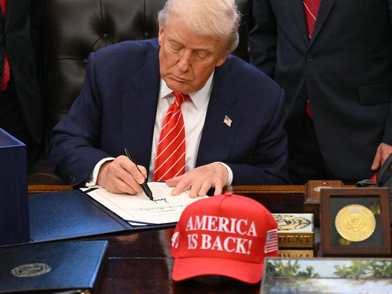 US-Präsident Trump unterschreibt ein Dokument im Weißen Haus. Vor ihm auf dem Schreibtisch eine Baseball-Mütze mit der Aufschrift "America is back" (Amerika ist zurück). Hinter Trump stehen mehrere Personen. US-Präsident Trump unterschreibt ein Dokument im Weißen Haus. Vor ihm auf dem Schreibtisch eine Baseball-Mütze mit der Aufschrift "America is back" (Amerika ist zurück). Hinter Trump stehen mehrere Personen.