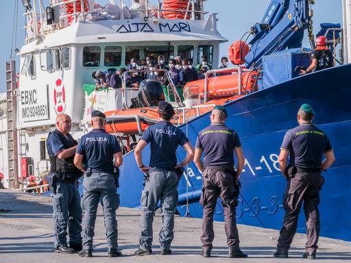 Auf einem Schiff im Hafen von Lampedusa warten viele Menschen darauf, an Land gehen zu können. Vor dem Schiff stehen fünf Polizisten.