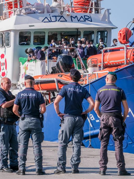 Auf einem Schiff im Hafen von Lampedusa warten viele Menschen darauf, an Land gehen zu können. Vor dem Schiff stehen fünf Polizisten.