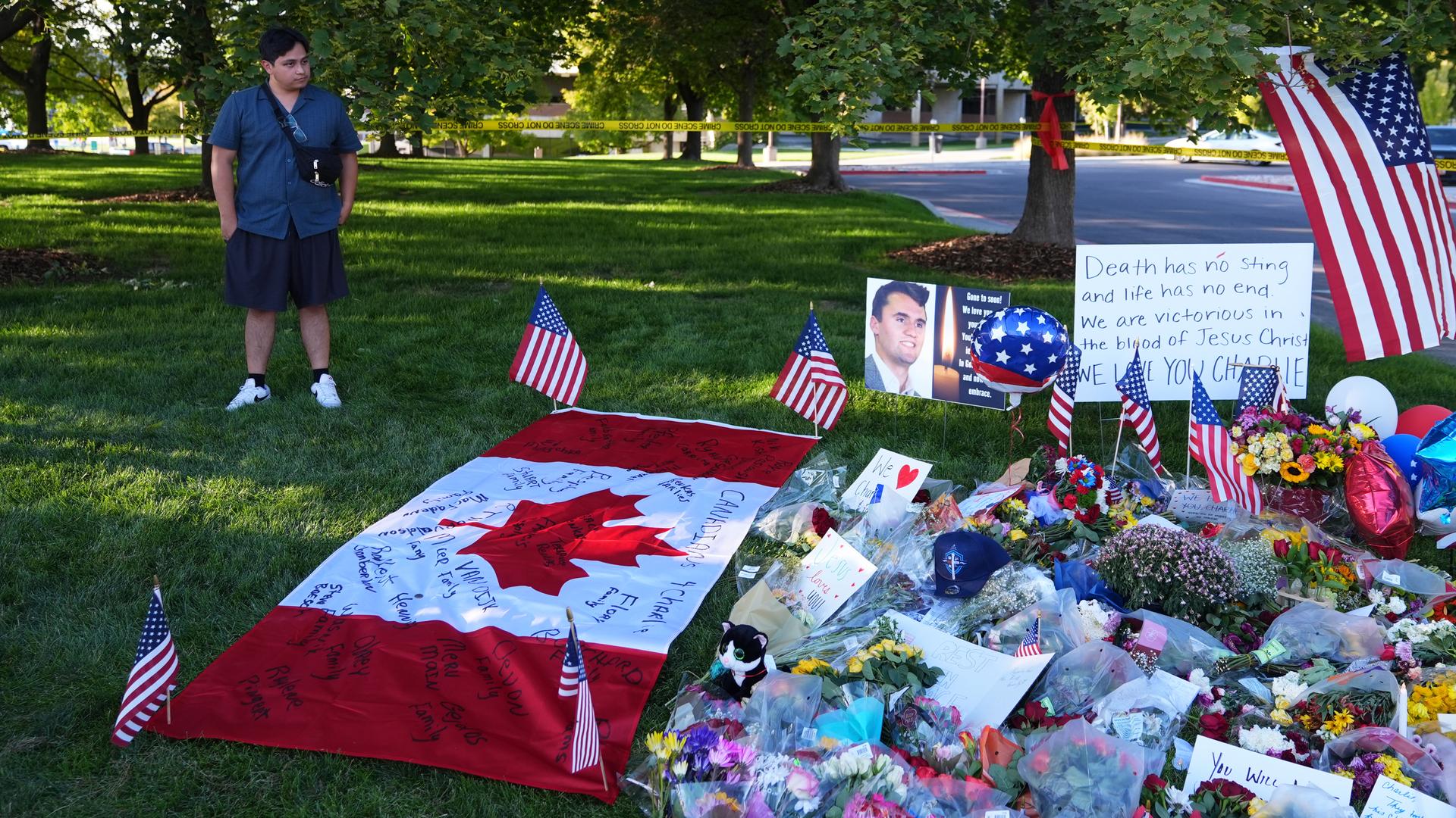 Ein junger Mann steht auf einer Wiese und blickt auf eine improvisierte Gedenkstätte für Charlie Kirk, wo Menschen Blumen, Bilder und Botschaften niedergelegt haben.