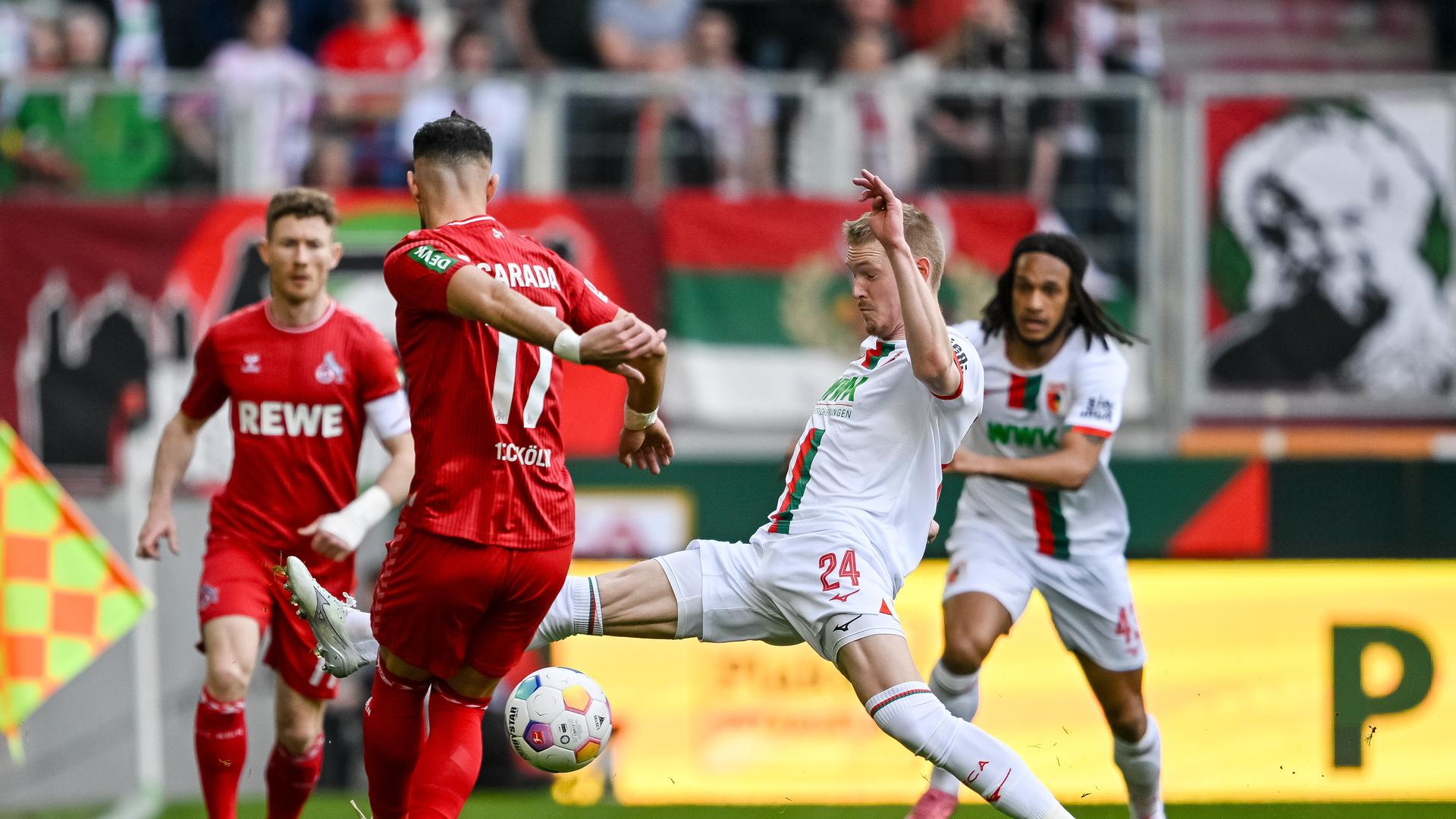 Das Foto zeigt die Kölner Spieler Florian Kainz und Leart Paqarada in Aktion gegen die Augsburger Fußballer Fredrik Jensen und Augsburgs Kevin Mbabu (l-r).