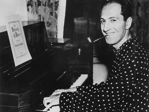 Ein Schwarzweiß-Foto zeigt den Komponisten George Gershwin am Flügel sitzend und in die Kamera lächelnd.