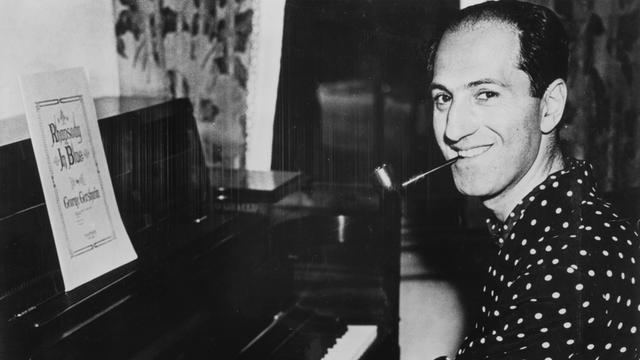 Ein Schwarzweiß-Foto zeigt den Komponisten George Gershwin am Flügel sitzend und in die Kamera lächelnd.